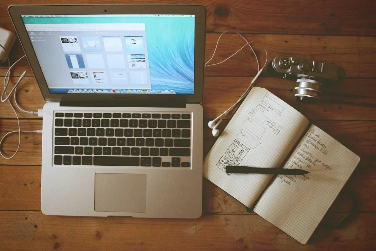 A laptop beside an open notebook with sketches on a wooden desk
